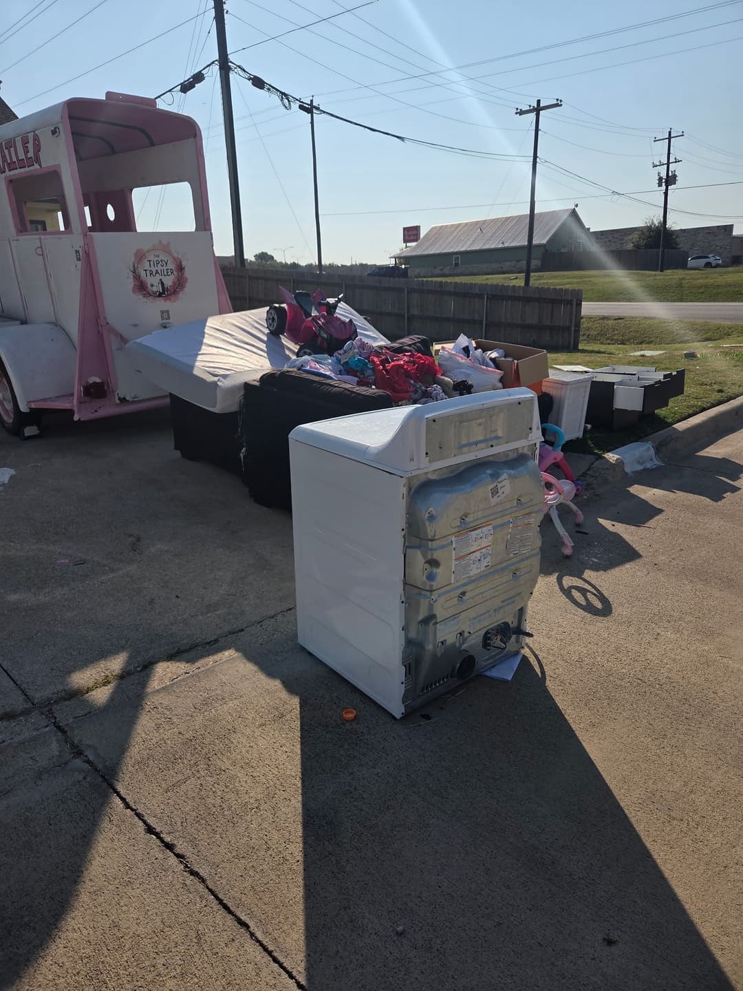 Abandoned washing machine among discarded furniture and bags near a roadside.