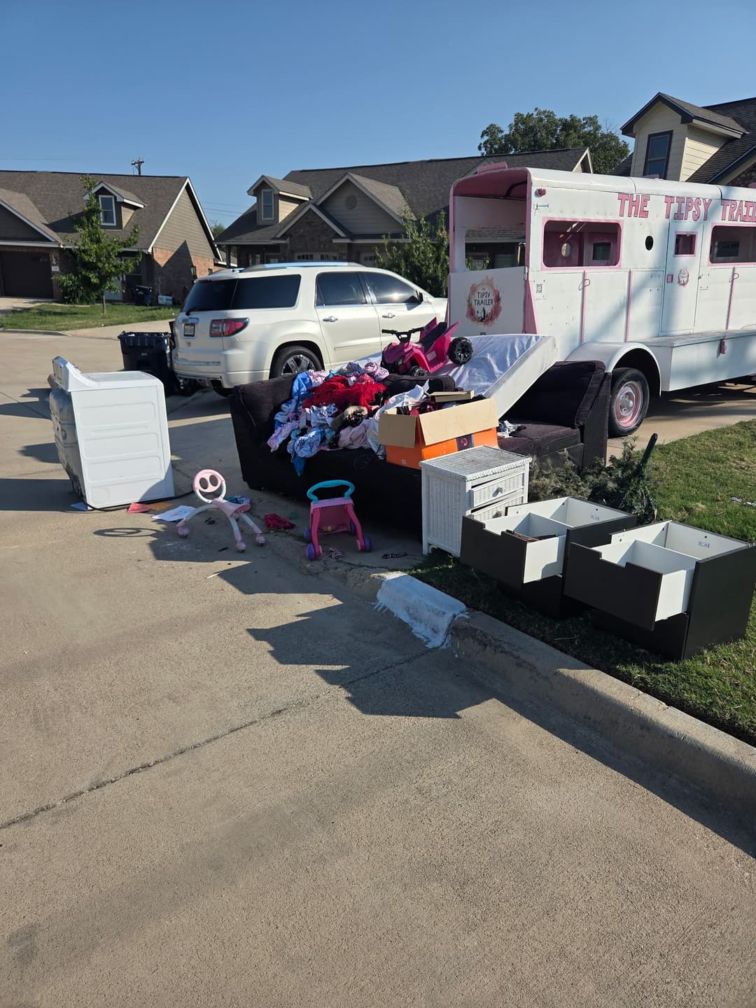 Piled clutter including furniture and boxes on curb near parked vehicle and trailer.