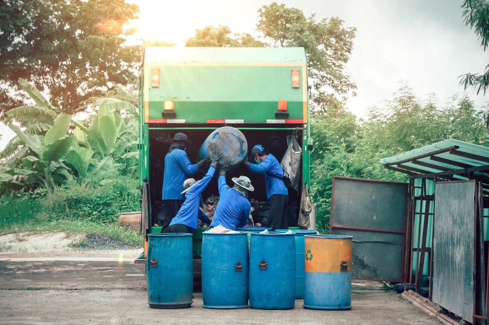 Waste collection workers loading trash into a green garbage truck beside blue barrels.